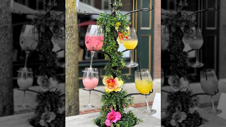 A metal stand holding four wine glasses,; two filled with pink liquid, two filled with yellow liquid decorated with greenery and pink and yellow flowers