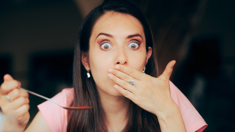A young woman with a spoon in her right hand appears to have just tasted something awful; her eyes are opened wide and her hand is over her mouth