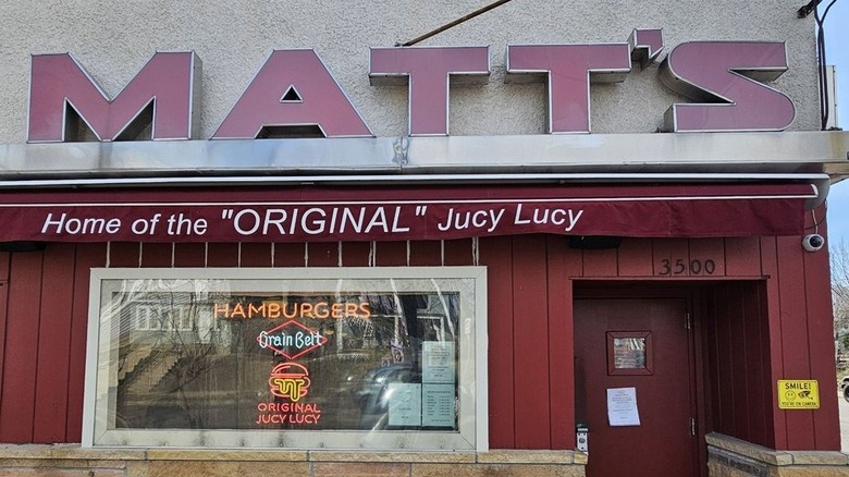 The exterior of Matt's Bar; "Matt's" reads in block letters against gray concrete; maroon awning reads "Home of the 'original' Jucy Lucy," with a window on the left with a neon sign in it and a door on the right