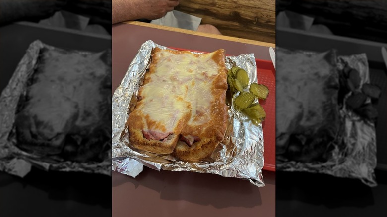 A red restaurant tray set on a brown surface; tray is lined with foil; two pieces of French bread, sliced lengthwise, sit on the foil; melted cheese tops the bread; 8 pickle slices are on the right