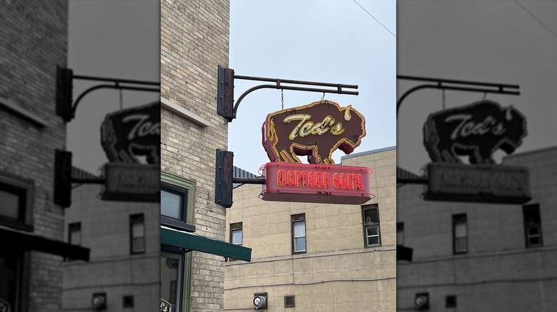 A vintage-style neon sign affixed to a light-colored brick building; sign is a brown bison with "Teds" in the center with "Montana Grill" in red below it
