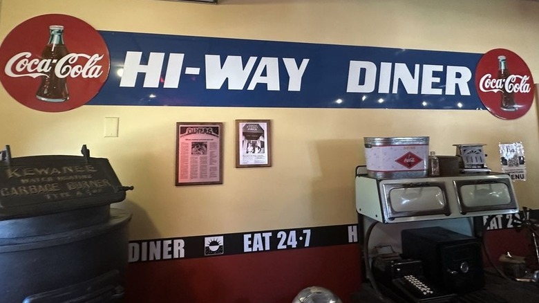 A butter yellow interior wall with a blue metal sign that has the words "Hi-Way Diner" writeen on it in white; sign is flanked by "Coca-Cola" signs; various vintage items appear below it