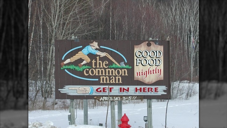 A brown roadside sign with winter woods in the background; sign depicts a farmer with a manual plow and reads "the common man," "good food nightly," and "get in here" with a hand pointing to the left