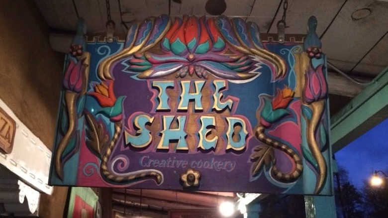 A sign for The Shed; "The Shed Creative Cookery" appears in stylized lettters in blue; sign is decorated with pink and red flowers with gold, purple, and teal flourishes