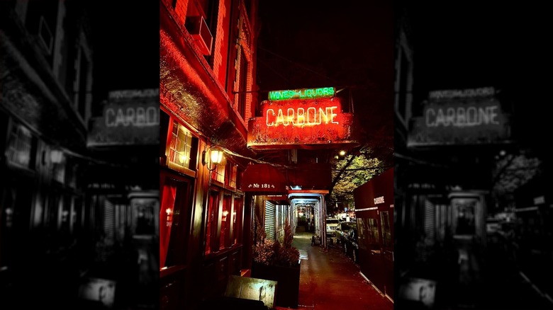 The neon sign for Carbone affixed to a brick building on a New York City street lit at night; "Carbone" appears in red; "Wines Liquors" appears in green above it