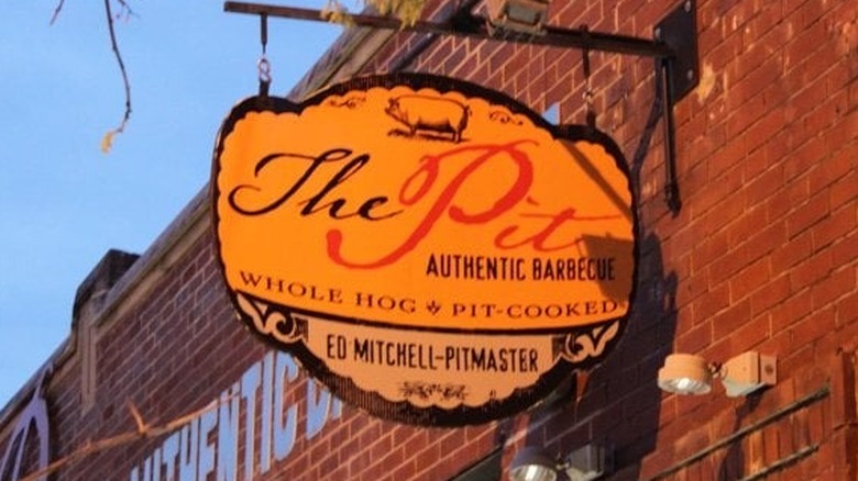 Yellow sign for The Pit affixed to a brown brick building; a small pig appears on top with "The Pit Authentic Barbecue, Whole Hog PIt-Cooked, and Ed Mitchell-Pitmaster" written on it; all letters are in black except "Pit," which is in orange