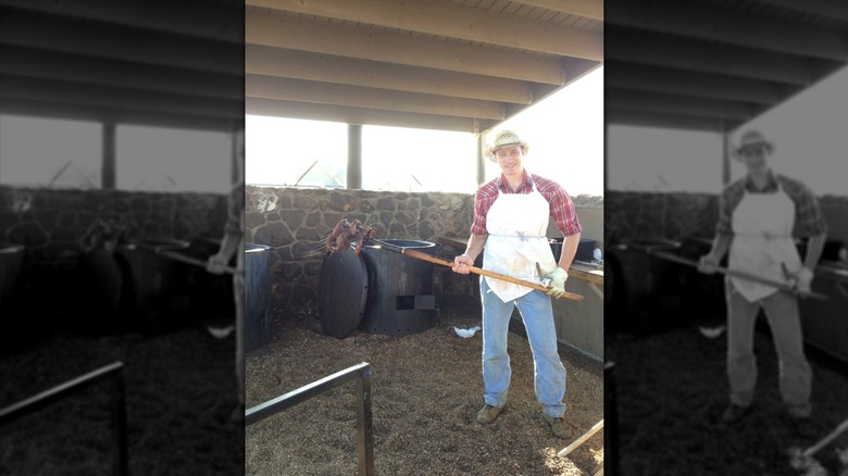 A man wearing a red flannel, jeans, a white aprong, and a cowboy hat standing in a covered outdoor area with stone walls; man is holding a pitchfork with two large steaks on it
