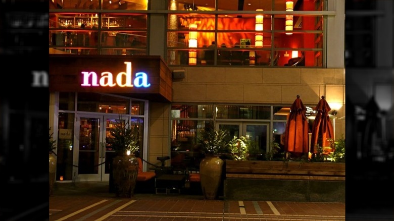 The exterior of a modern city building at night; the word "nada" is lit in pink, yellow, yellow, and blue letters, respectively; double-door entrance on left; windows on the right