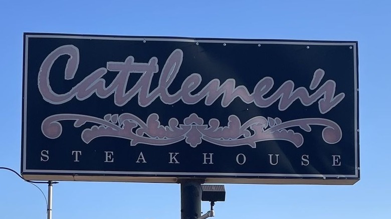 The outdoor sign for Cattlemen's Steakhouse. "Cattlemen's" is in gray cursive on a dark blue background with a flourish underneath with "steakhouse" in Roman capital letters