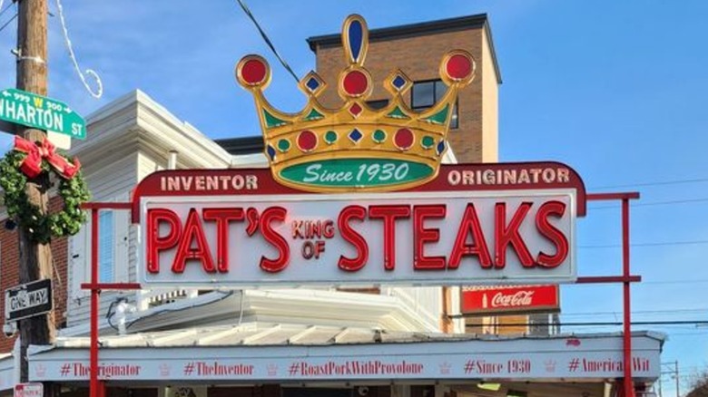 The exterior sign for Pat's King of Steaks in daylight: "Pat's King of Steaks" is in red on a white background with a gold crown set above it and "Inventor, Originator" above the restaurant name and "Since 1930" written in the crown