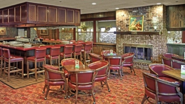 The lounge at Twin Oaks; dated brown and red patterend carpeting, maroon club chairs surround 4-top tables with a rectangular-shaped bar in the background;  a brick fireplace, wood paneling, and windows line the walls