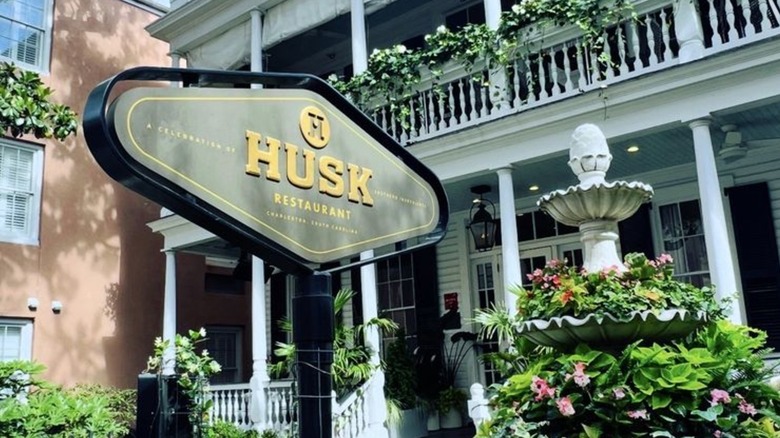 The exterior sign for Husk: Husk Restaurant is written in gol don a gray background; a white, two-story antebellum-style building decorated with greenery is in the background
