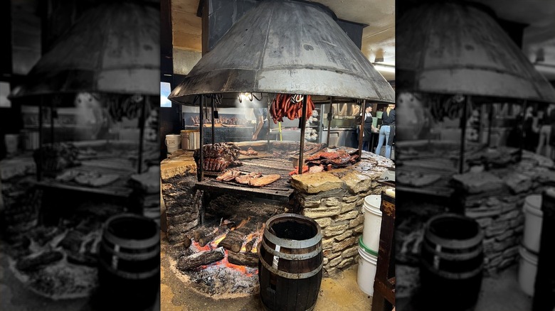 A large brick indoor barbecue pit with a sheet metal hood; various meats are on the grill and sausages hang above it