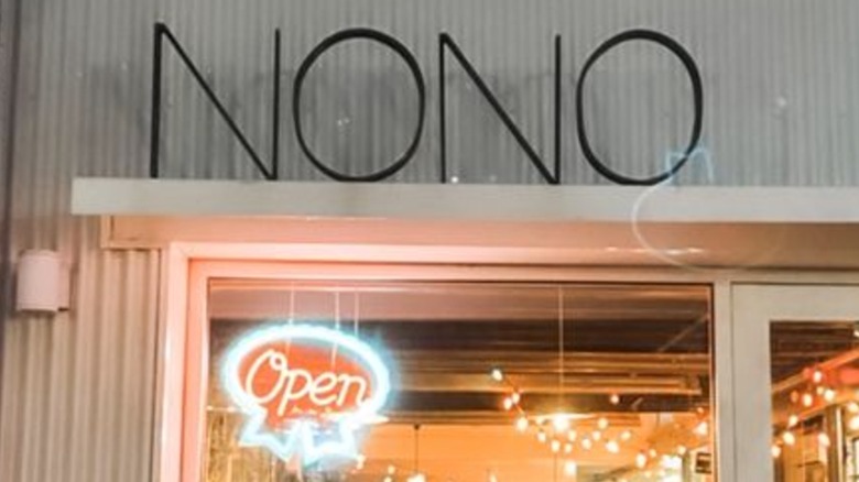 The exterior. sign for Nono: "Nono" in thin metal letters against gray metal paneling set above a window with a neon sign that says "open" in it