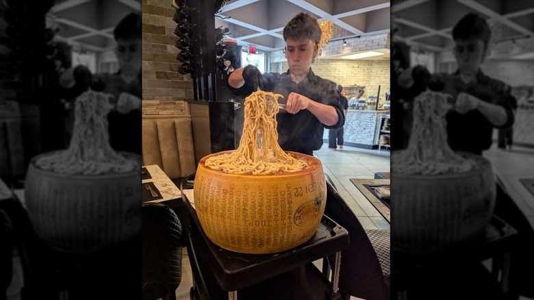 A young male server in a black shirt tossing spaghetti-shaped pasta atop a large wheel of pecorino romano
