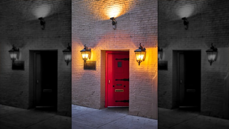 The exterior of a cream-colored brick building at night: a red door is lit by a light above and one on each side