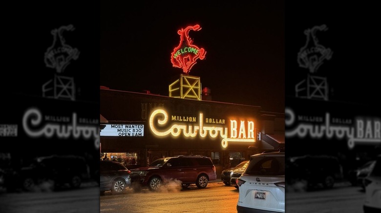 The exterior neon sign for Million Dollar Cowboy Bar at night: "Million Dollar Cowboy Bar" is affixed to the single-story building in yellow neon; a stylized cowboy on a bucking bronco in red neon with "welcome" written in green above it