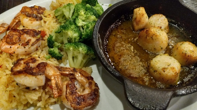 A plate of Ruby Tuesday's Shrimp and Scallop meal, with shrimp on a bed or rice and broccoli next to a pan full of seared scallops.