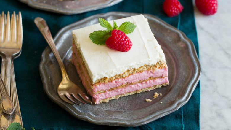 Icebox cake piece with pink raspberry filling topped with white cream, a raspberry, and a mint sprig on a metal plate with a fork