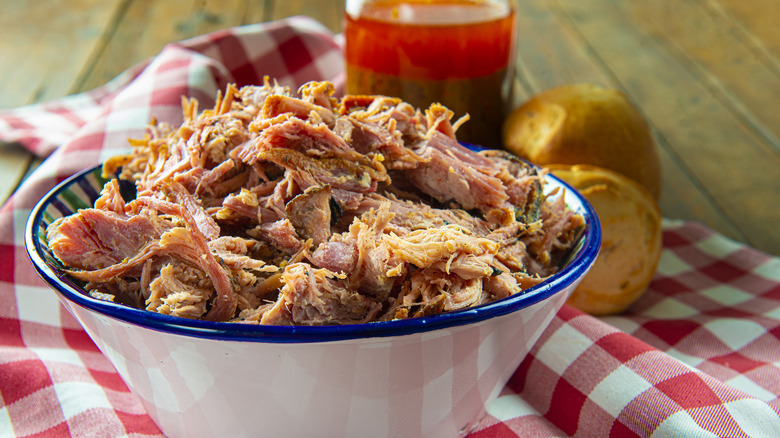 A white bowled filled with pulled pork on top of a white and red checkered table cloth with a container of vinegar barbecue sauce in the background.