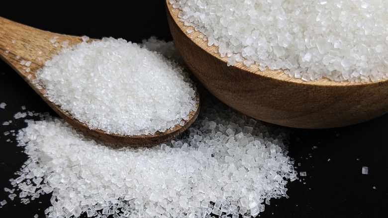 Granulated white sugar spilling out of a wooden bowl onto a wooden spoon.