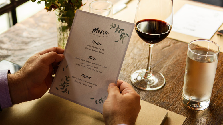 A man holds a paper menu at a restaurant table with a glass of wine and water