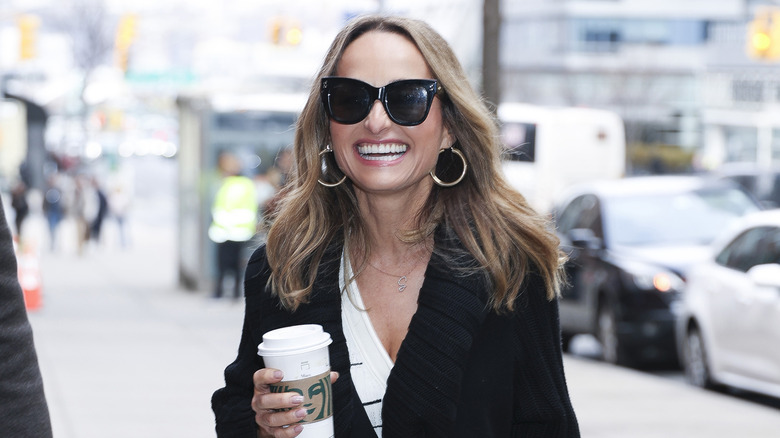 Giada de Laurentiis wearing big black sunglasses and smiling while holding a Starbucks coffee outdoors