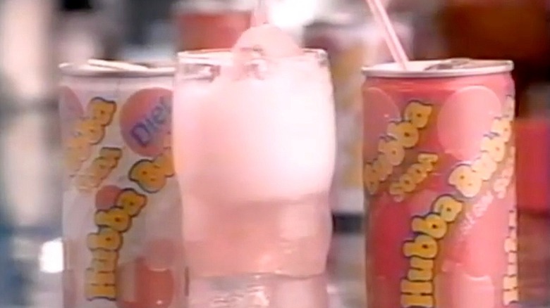 A still from a 1988 Hubba Bubba soda commercial showing the pink and yellow can sitting next to a glass full of the pale pink soda and a can of the diet version