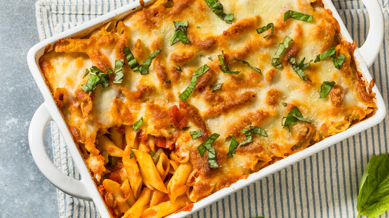 Close up of a white casserole dish filled with cheese-topped baked pasta garnished with fresh basil