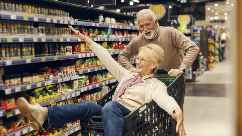 Two grandparents goofing off in a grocery store by riding in the shopping cart with their arms out like an airplane.