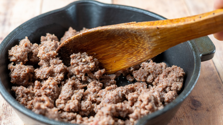 A wooden spoon stirring a cast iron skillet of cooked ground beef.