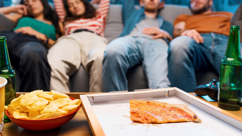 Tired adults laying on couch with pizza and chips on table