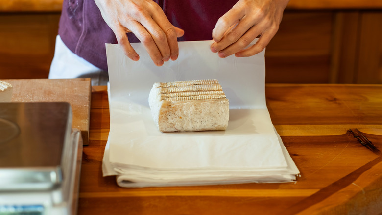 A person wraps cheese in paper on a wooden counter