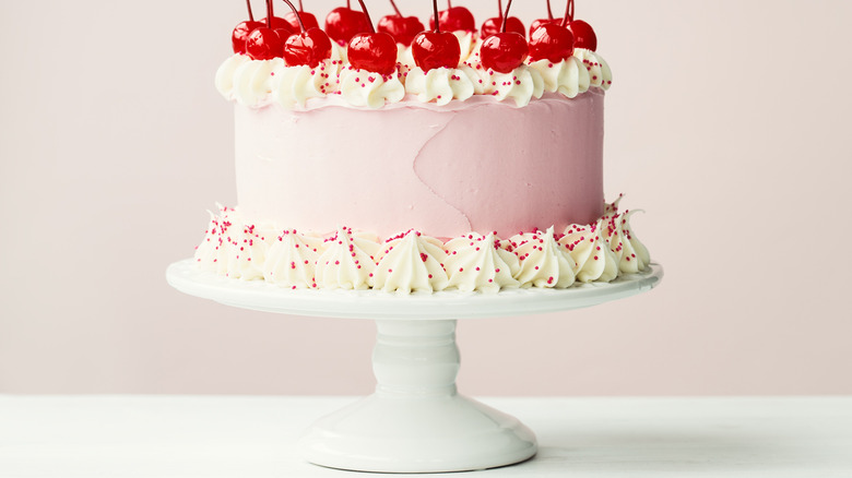 Pink cake on stand decorated with whipped cream, maraschino cherries