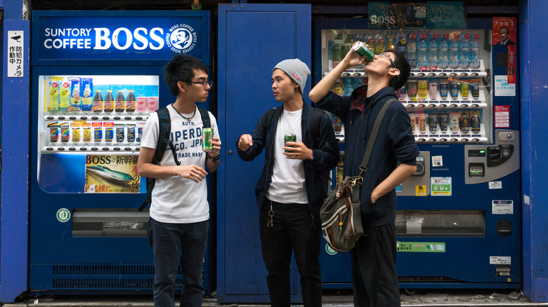 Three young people drinking canned beverages in front of vending machines