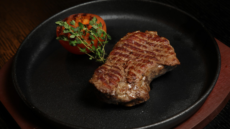 A delmonico steak in a pan beside a grilled tomato and herbs