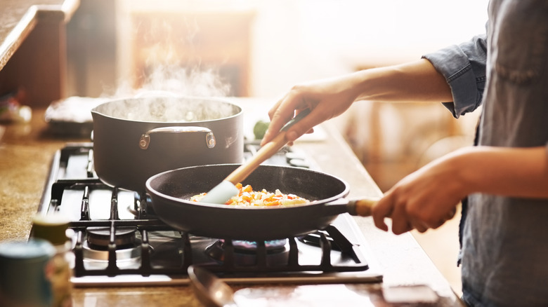 Person cooking at stove with hands handling a pan of food and a steaming pot next to it