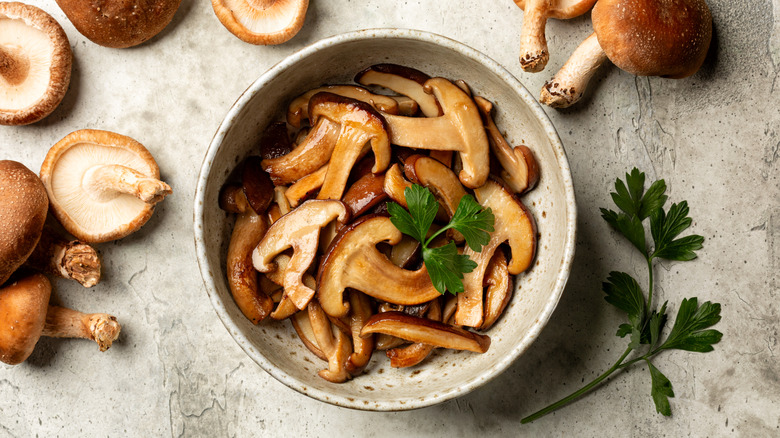 Top view of a bowl of cooked mushrooms surrounded by raw mushrooms