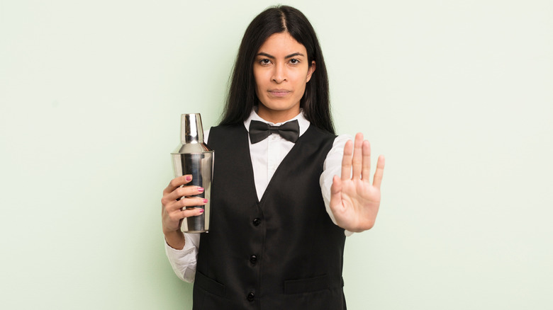 A young woman in a bartender's uniform raises a hand in a "stop" gesture while holding a shaker