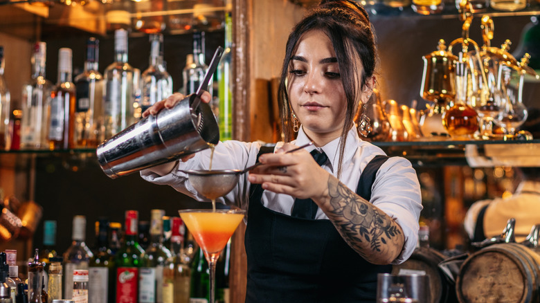 A bartender with floral tattoos straining an orange drink into a glass