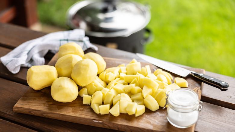 Potatoes cut into chunks with a chef's knife on a cutting board and wooden table, outside with grass blurred in the background.