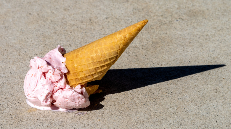 A scoop of strawberry ice cream in a waffle cone has fallen on the ground and is melting.