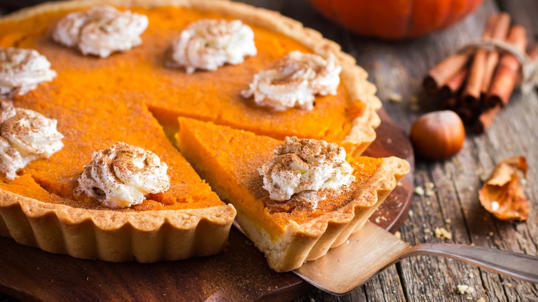Person taking slice of pumpkin pie with dollops of whipped cream topped with cinnamon on wood surface