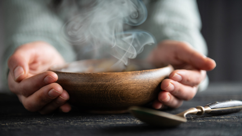 A pair of hands holding steaming wooden bowl of soup, with a spoon in the foreground