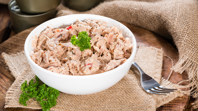 A bowl of tuna salad topped with a sprig of parsley, surrounded by burlap mats and cans of tuna