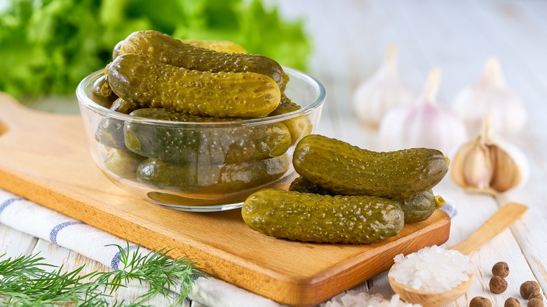 Pickles in a small round glass bowl surround by heads of garlic, dill, and a spoon with large salt crystals