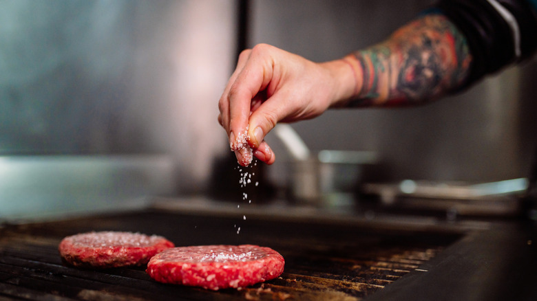 A hand sprinkling salt onto cooking burgers.