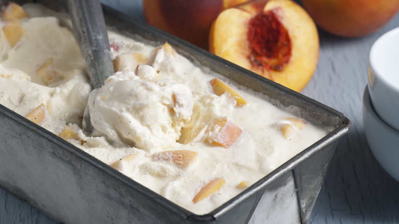 Metal loaf pan filled with peach ice cream with an ice cream scoop inside -- cut peaches are in  the background