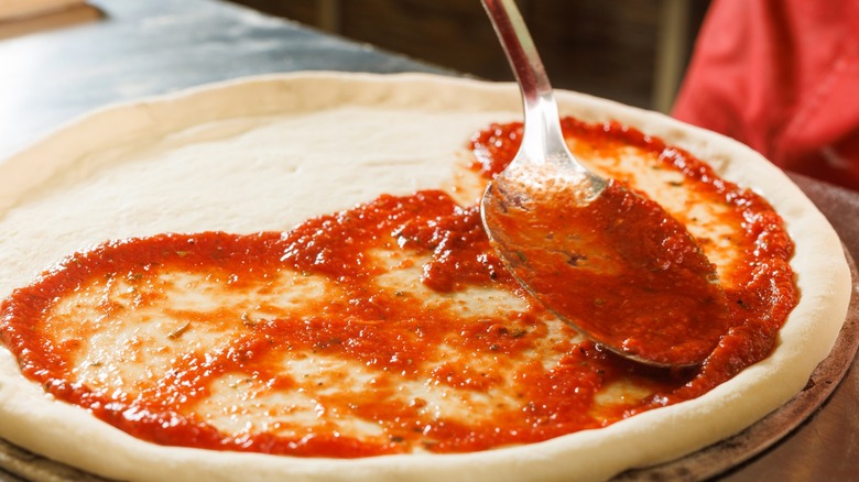 Large metal spoon spreading tomato sauce on pizza dough