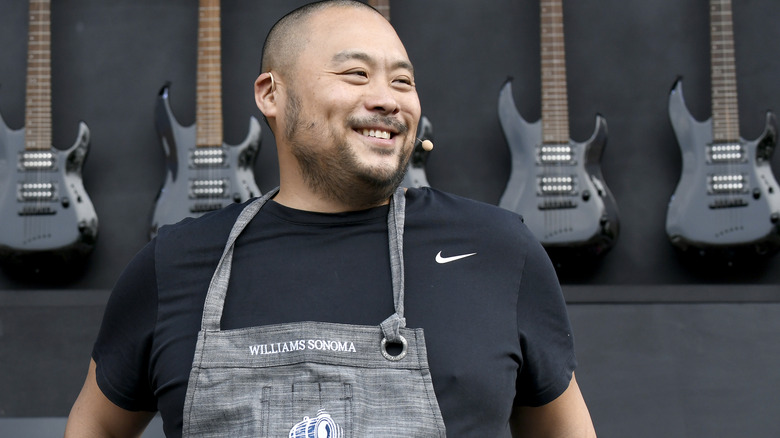 Chef David Chang smiling during a cooking demonstration with guitars hanging behind him
